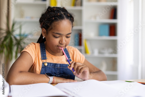 Slika na platnu Black teenage girl chewing on pencil while concentrating on her notebook, doing
