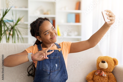 Bild auf Leinwand Black teenage girl taking selfie on smartphone and showing peace sign while sitting at home