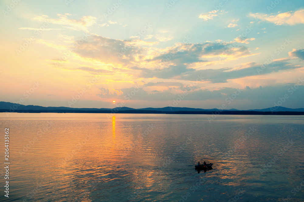 Obraz premium Summer vacation landscape, the sun setting behind the mountains and water surface lit by sunset and an unidentified people on a boat at the distance