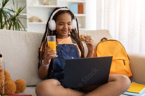 Schilderij op canvas Black teenage girl enjoying cookie and orange juice while using laptop at home
