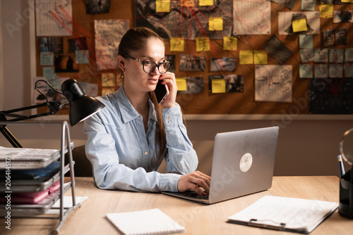 Foto A Caucasian female detective sits at a table in front of an evidence board, focused on working on a laptop while talking on a smartphone