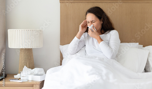 Foto Sick woman sneezing into a tissue while sitting in bed, ill adult female showing