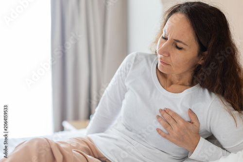 Fotografie Woman sits on bed holding hand to chest with pained expression, chest pain or he