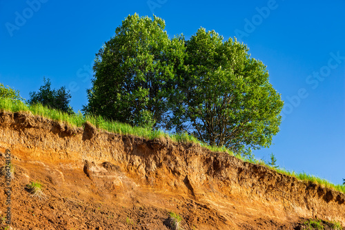Steep sandy quarry slopes topped with lush green grass and vibrant trees against a deep blue sky, low angle shot capturing raw geological layers and natural summer vegetation