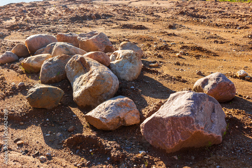 Large granite boulders gathered together in a sandy quarry under bright sunlight, raw natural stones piled on rough ground creating a rustic texture for construction or earthworks
