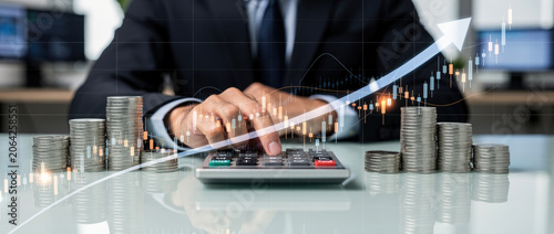 Businessman calculating financial growth with stacks of coins and upward trending graph overlay.