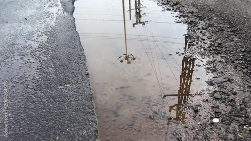 Reflections of electric poles, power lines, and trees shimmering in rainwater collected in a roadside pothole, creating an abstract urban landscape after rainfall
