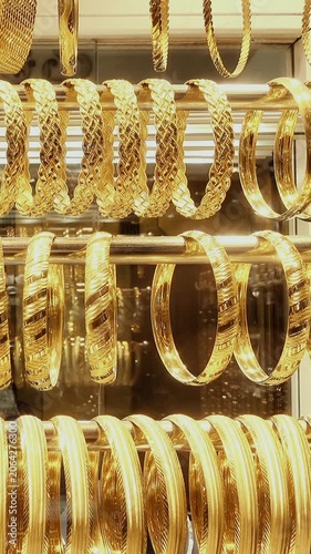 Rows of shiny gold bangles and bracelets hang in a jewelry shop display case as the camera holds on the ornate metal designs and reflections