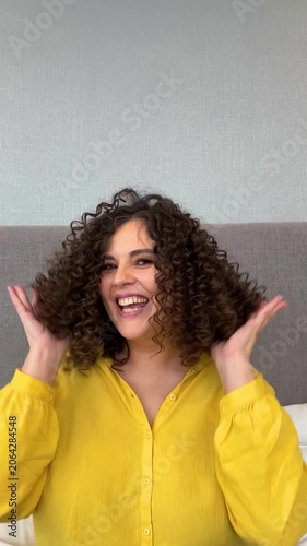 Vertical 4K video. 60 fps. Young adult white child free woman with curls, yellow shirt in hotel room smiles, touches her bouncing curly hair looks at camera, laughing. Diverse beauty, leisure concept