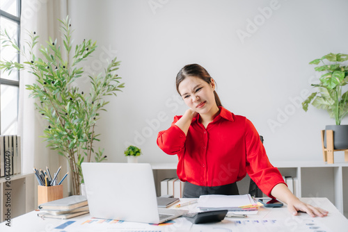 Schilderij op canvas Desk Discomfort: A businesswoman in a red shirt experiences neck pain while working at her office desk, highlighting the physical toll of prolonged computer use