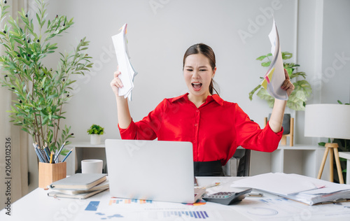 Photos Frustrated Professional Amidst Chaos: A woman in a red shirt sits at her desk, overwhelmed by papers, her hands raised in exasperation while working in an office