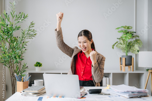Obraz na płótnie Excited businesswoman celebrating success at her desk: A young, energetic businesswoman with her fist raised in triumph, beaming with joy as she looks at her laptop screen in a modern office setting