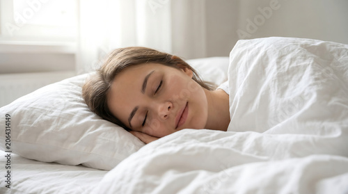 Woman sleeps peacefully in a soft white bed under natural morning light that fills the bedroom with a calm and serene atmosphere.