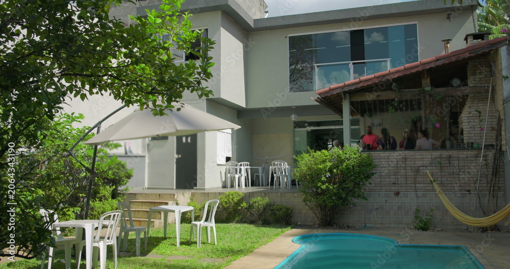 Obraz premium Sunny backyard scene with umbrella-shaded tables, hammock, swimming pool, and group socializing at covered grill area of modern suburban home