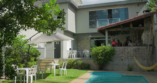 Sunny backyard scene with umbrella-shaded tables, hammock, swimming pool, and group socializing at covered grill area of modern suburban home
