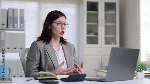 Professional woman with glasses sitting at a desk and having a video conference on her laptop, gesturing while talking during an online meeting in a bright corporate office environment