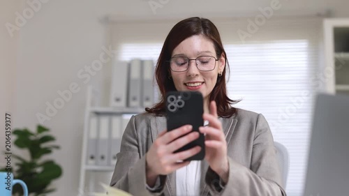 Smiling businesswoman in eyeglasses enjoying a break from work, sitting at her desk and scrolling through her smartphone, engaging with social media, texting friends, or reading news online