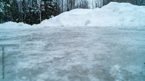 Fotografie A dramatic low-angle ground-level photograph looking across a thick, textured sheet of frozen glare ice toward large plowed snowbanks and winter forest trees on a cold, overcast day