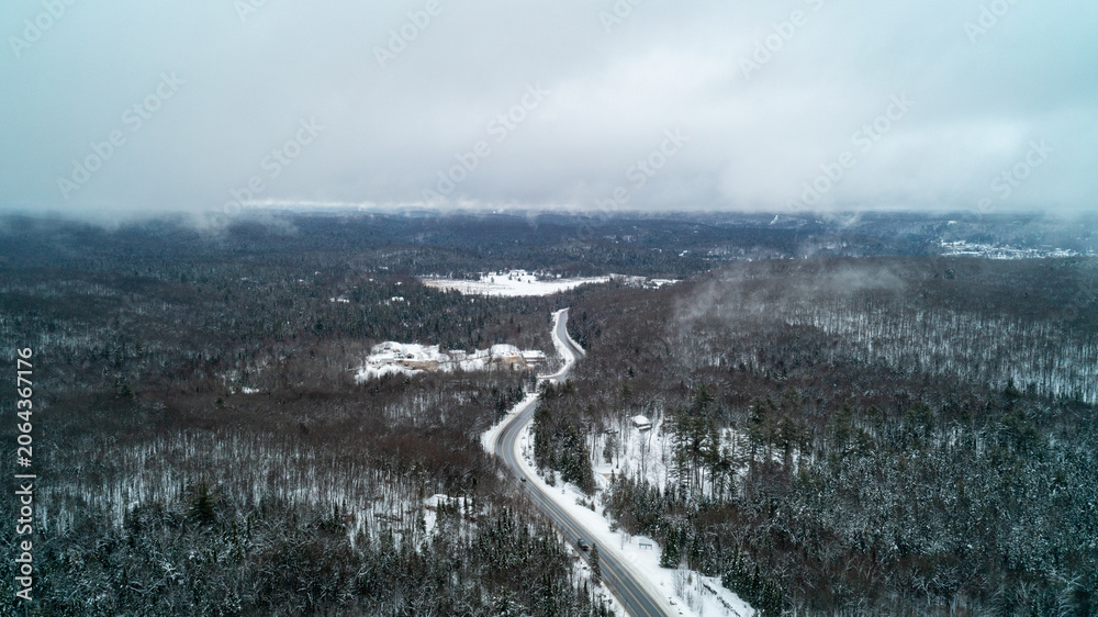 Fototapeta premium A sweeping high-angle drone photograph over a winding two-lane highway cutting through a dense winter forest, with low hanging fog and mist drifting below a moody, overcast sky.
