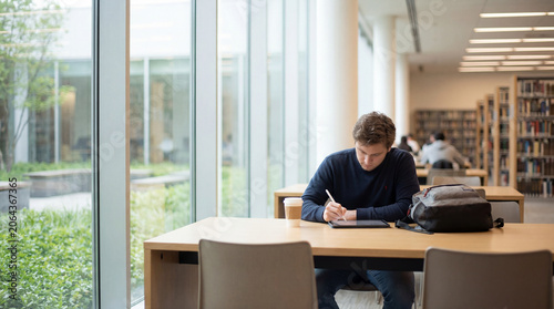 Young student using stylus on digital tablet beside coffee backpack in modern library with window light showing focused study academic research campus lifestyle quiet concentration