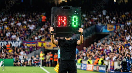 Soccer referee holding up digital substitution board displaying numbers fourteen and eight in front of crowded stadium during football match.