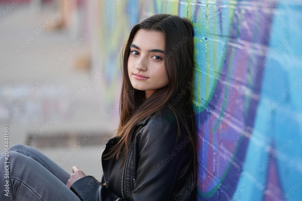 Obraz premium Teenage girl leaning against colorful graffiti wall wearing black leather jacket