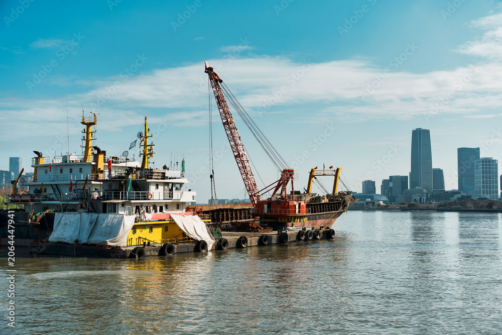 Fototapeta premium Floating Crane Barge and Cargo Ship on River With City Skyline Background