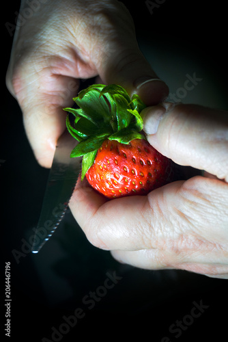 Plano de detalle de manos de persona mayor pelando o cortando una fresa roja brillante con un cuchillo de cocina pequeño. Iluminación dramática estilo clave baja sobre fondo negro.