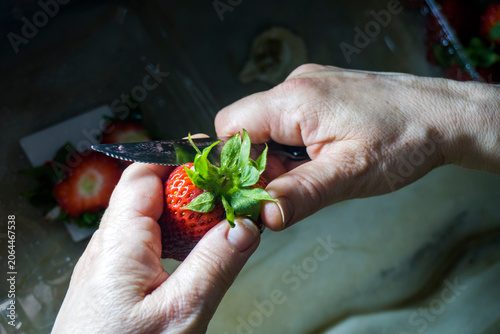 Primer plano de manos de un adulto mayor limpiando y cortando el pedúnculo de una fresa roja fresca con un cuchillo de cocina bajo iluminación de alto contraste y estilo dramático.