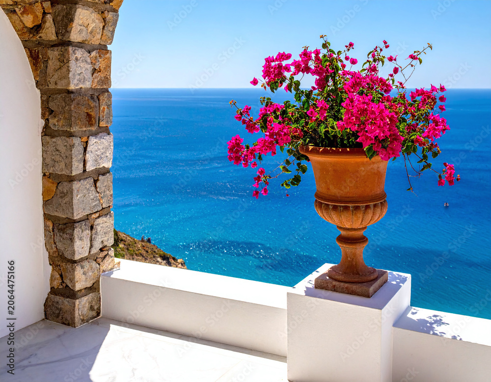 Fototapeta premium Vibrant pink bougainvillea flowers in a classic terracotta urn sit on a white balcony, overlooking the breathtaking, peaceful turquoise Mediterranean Sea on a sunny summer day.