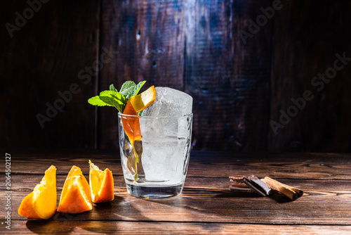 Whiskey glass with ice cubes, mint and orange peel, orange slices, cinnamon sticks and chocolate on dark low key spotlit wooden background.