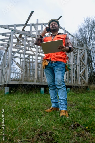 Modern construction manager coordinating logistics via mobile phone in front of a wooden building structure