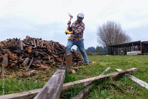 Mid-adult lumberjack splitting logs with an axe next to a large pile of wood