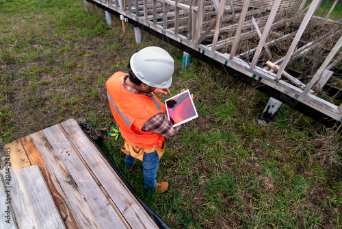 Builder in hard hat and safety vest reviewing architectural plans on a screen near wood materials
