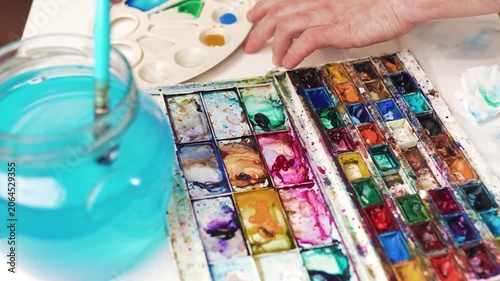 Paintbrush dipping into green watercolor cuvette with water jar beside