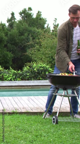 Vertical video: Cooking man in olive top with green bottle and tongs at grill serving friend coming