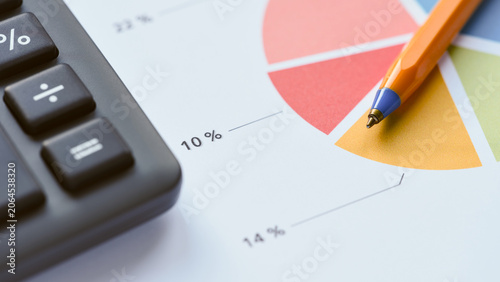 An orange pen lying on a colorful pie chart document next to a black office calculator, symbolizing financial planning and business analysis. Close up.