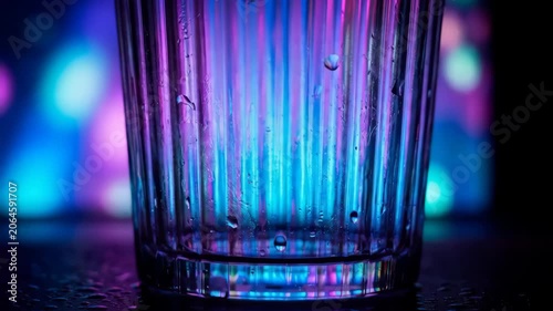 Close-up of a transparent drinking glass illuminated by vibrant neon lights. Abstract background featuring colorful reflections, glass texture, moisture droplets, and modern nightlife aesthetics.