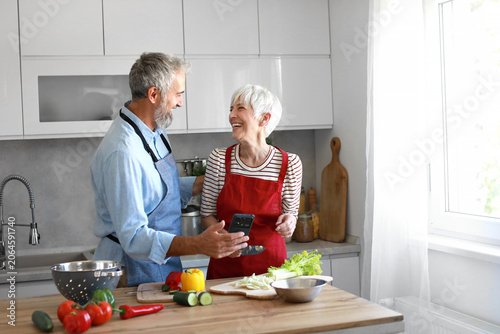 Portrait of happy senior mid aged mature couple prepering meal with fresh vegatebles and following internet instructions for a recipe on a smartphone or looking at mobile phone message or website app 