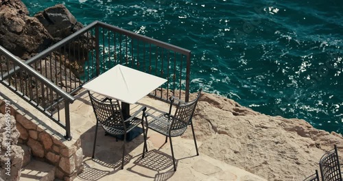 A table and chairs are positioned on a rocky waterfront terrace above the Adriatic Sea near Primosten, Croatia.