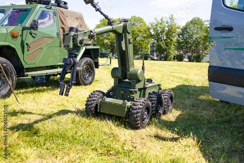 A large wheeled unmanned ground vehicle with a heavy robotic manipulator arm parked on green grass near a military armored truck on a sunny day.