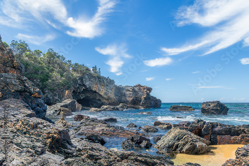 A sea cave in a headland and a shore lined with geologically interesting jagged rocks under a beautiful sky show some evidence of a tsunami in the past at Mystery Bay in New South Wales, Australia.