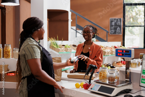 Obraz na płótnie African American vendor serves a customer at a checkout counter using a debit card for wireless payment at eco friendly local store; joyful client making cashless transactions at cashier desk