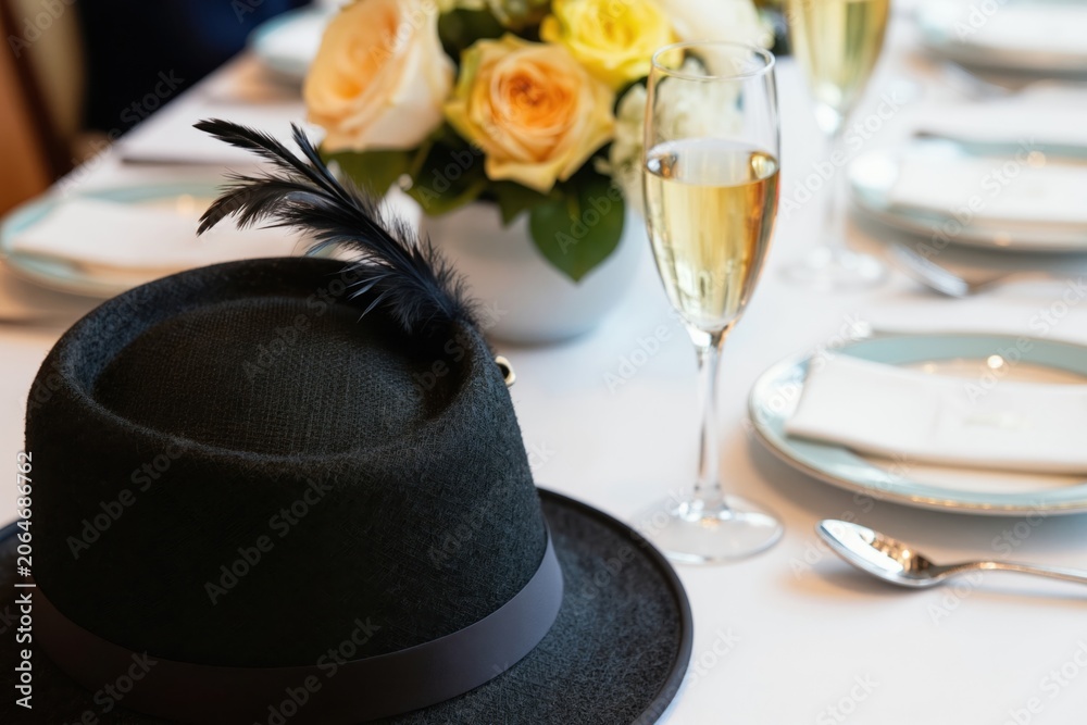 Fototapeta premium Elegant table setting for horse race event with champagne, flowers and formal hat. Guests enjoy celebration with fine china plates and glassware. Sophisticated festive gathering in soft light.