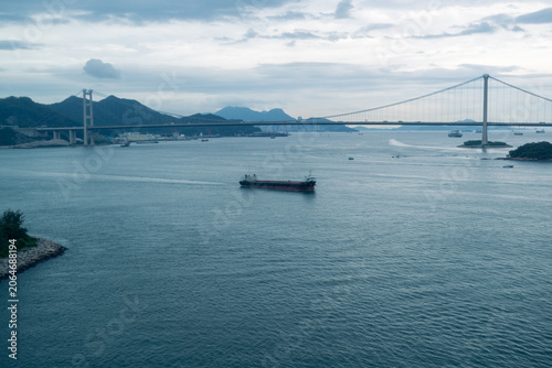 Cargo ship sailing under suspension bridge over calm sea during overcast day with distant mountains