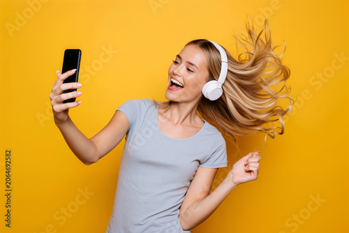 Young woman with flowing hair listens to music on headphones and takes a joyful selfie, framed by a vibrant yellow background. Generative AI.