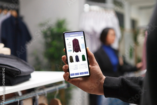 Tableau sur toile Caucasian customer in a clothes store is holding a smartphone and looking at clothes on a boutique website, male shopaholic is perusing clothing items in a mall using a cellphone