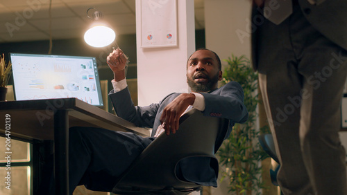 Cuadro en lienzo African American hyperactive employee sneaking up on tired coworker asleep in desk chair during night shift, happy businessman shaking up sleepy colleague taking nap in office
