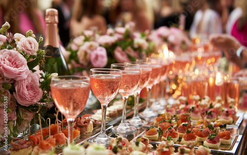 Row of pink cocktails on bar with appetizers and floral decor. Guests mingle at elegant event. Champagne bottle, food, drinks, and flowers create festive atmosphere