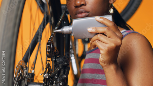 Tablou pe pânză BIPOC technician using specialized lubricant to repair squeaky bicycle chain, orange studio background, specialist applying oil on noisy bike parts during maintenance process, close-up shot
